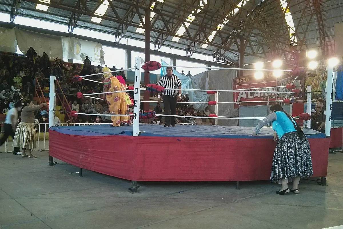 A wrestling ring with a red apron in a large covered hall, with a referee in the ring and spectators in the stands behind