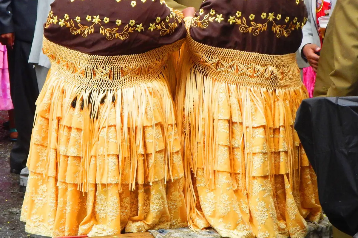 Two women seen from behind in layered yellow polleras with fringed, embroidered brown shawls at a festival in La Paz