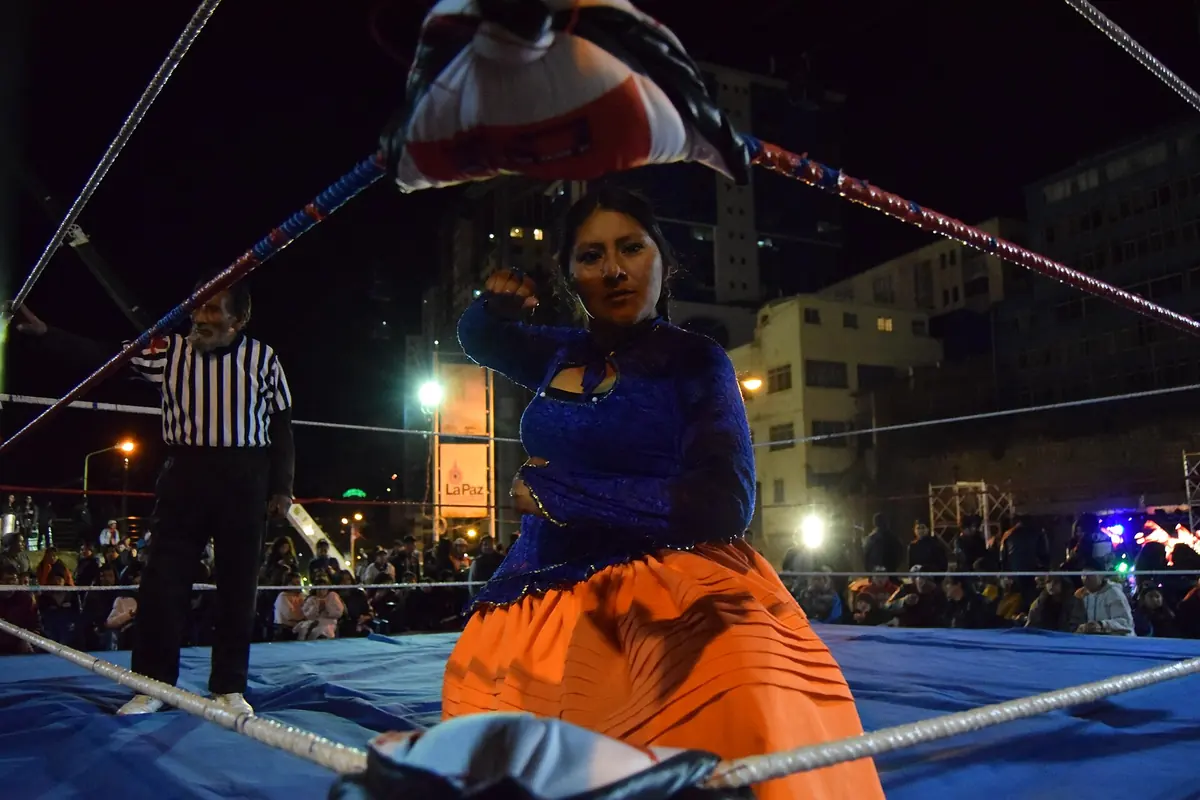 A cholita in a blue top and orange pollera kneeling on the mat near the ropes, a referee in a striped shirt behind her